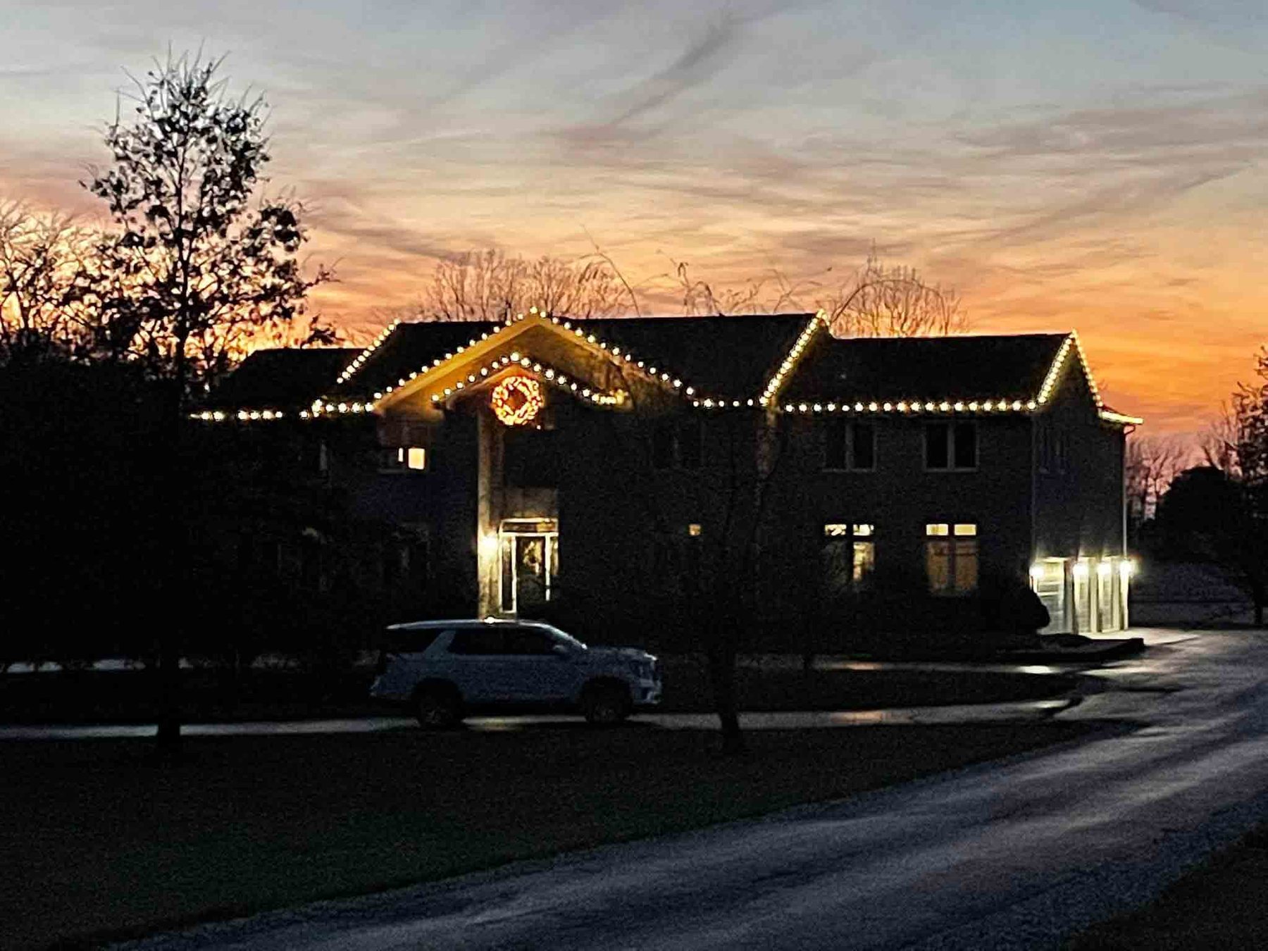 Large Indiana home lit with warm white Christmas lights along every roofline at sunset, with a lit wreath over the entrance